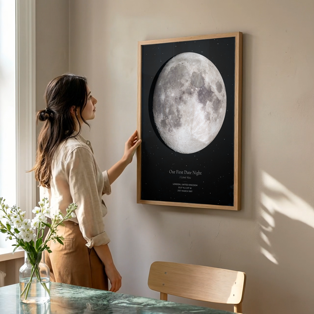 A woman in a beige blouse and brown pants stands beside a framed wall print showing a large moon on a black background with the text 'One Full Star Night' and 'Earth You.' The room has warm neutral walls, a wooden chair, and a vase of white flowers in the foreground.