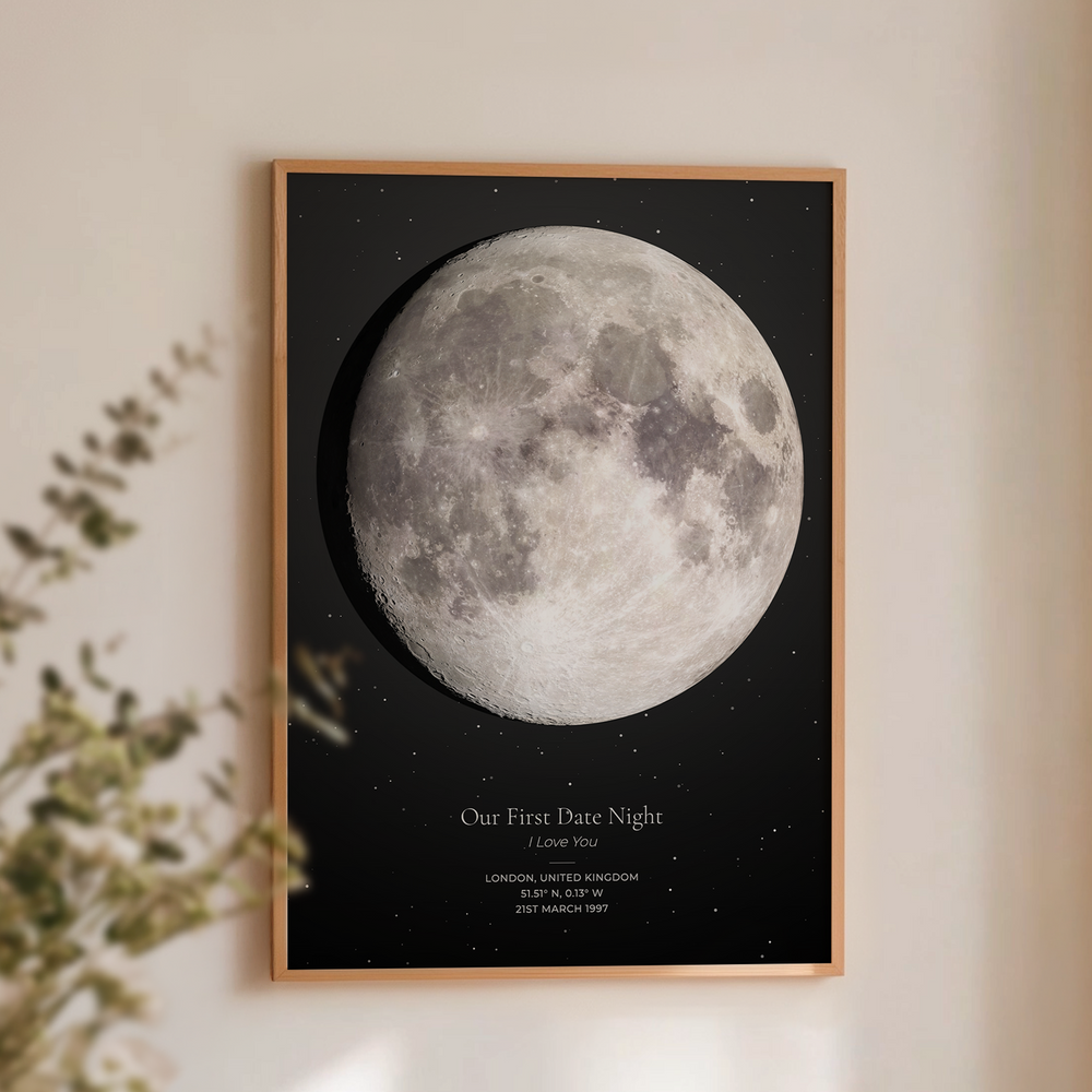 A framed custom moon print featuring a detailed grayscale moon on a black starry background, with the text 'Our First Date Night I Love You' and location/date details. The print is framed in a light wood frame and displayed on a warm beige wall with soft shadows and a blurred plant in the foreground.