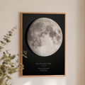 A framed custom moon print featuring a detailed grayscale moon on a black starry background, with the text 'Our First Date Night I Love You' and location/date details. The print is framed in a light wood frame and displayed on a warm beige wall with soft shadows and a blurred plant in the foreground.