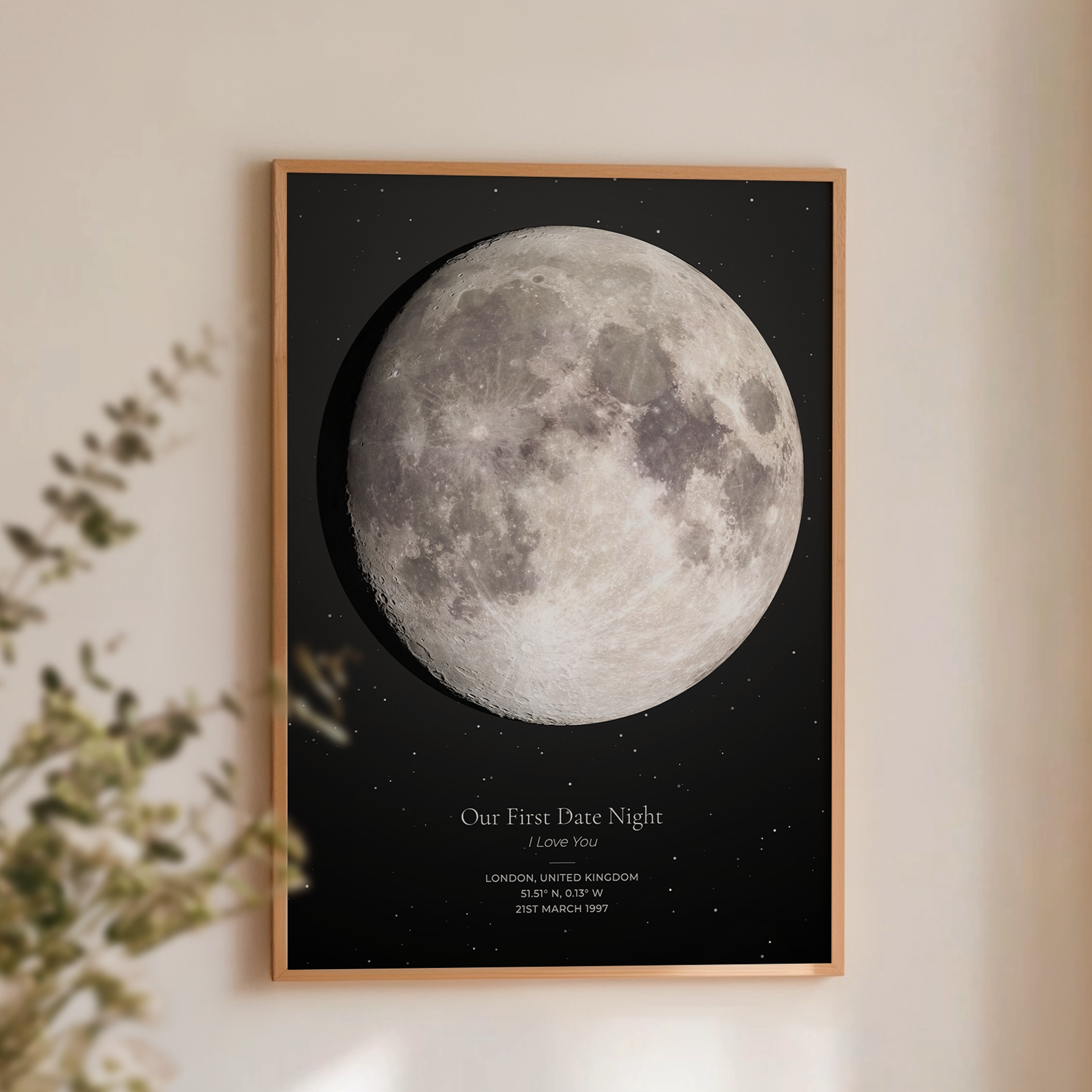 A framed custom moon print featuring a detailed grayscale moon on a black starry background, with the text 'Our First Date Night I Love You' and location/date details. The print is framed in a light wood frame and displayed on a warm beige wall with soft shadows and a blurred plant in the foreground.
